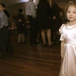 A little girl in a white dress standing in a room full of people.