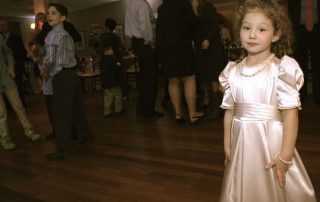 A little girl in a white dress standing in a room full of people.
