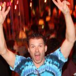 A man in a tie dye shirt with his hands up at a party.